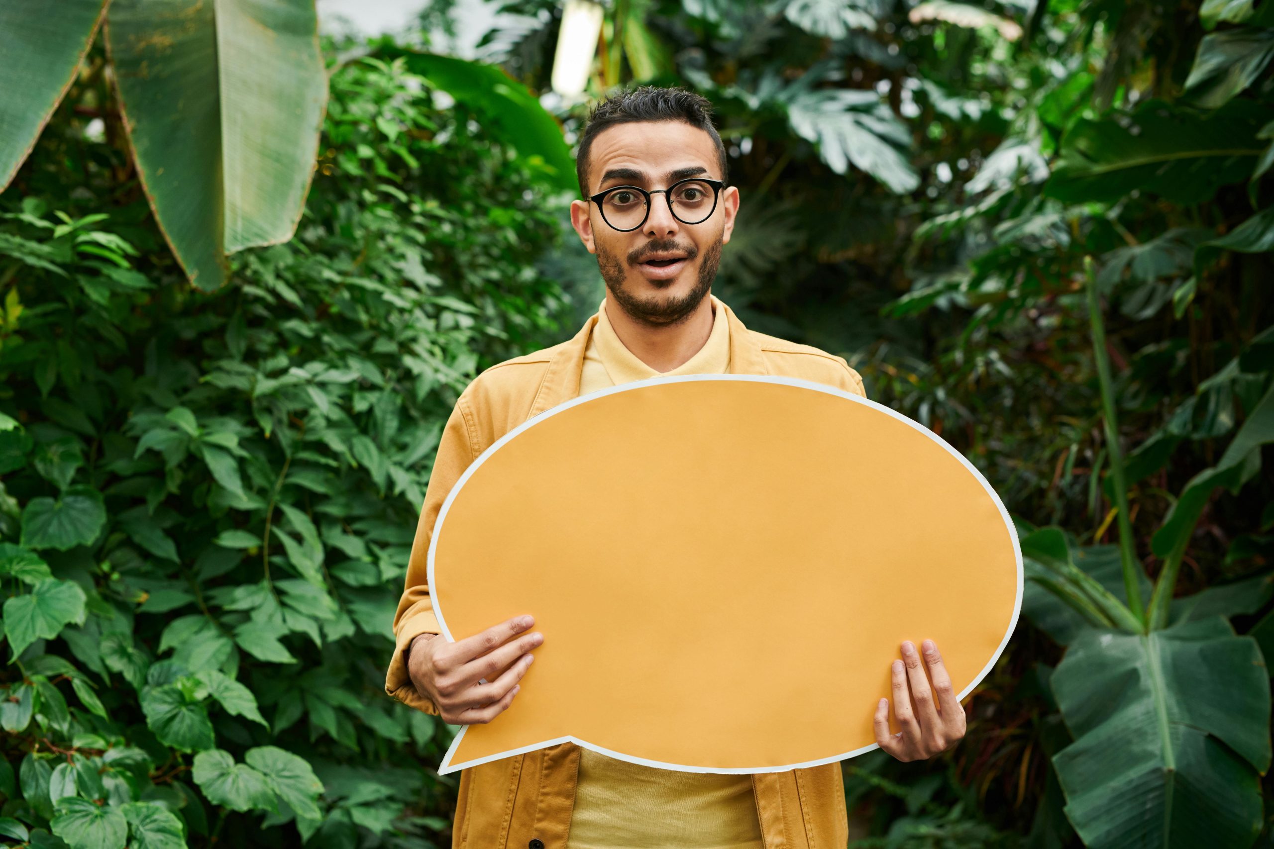 Surprised man with glasses holding a yellow speech bubble in lush green garden setting.