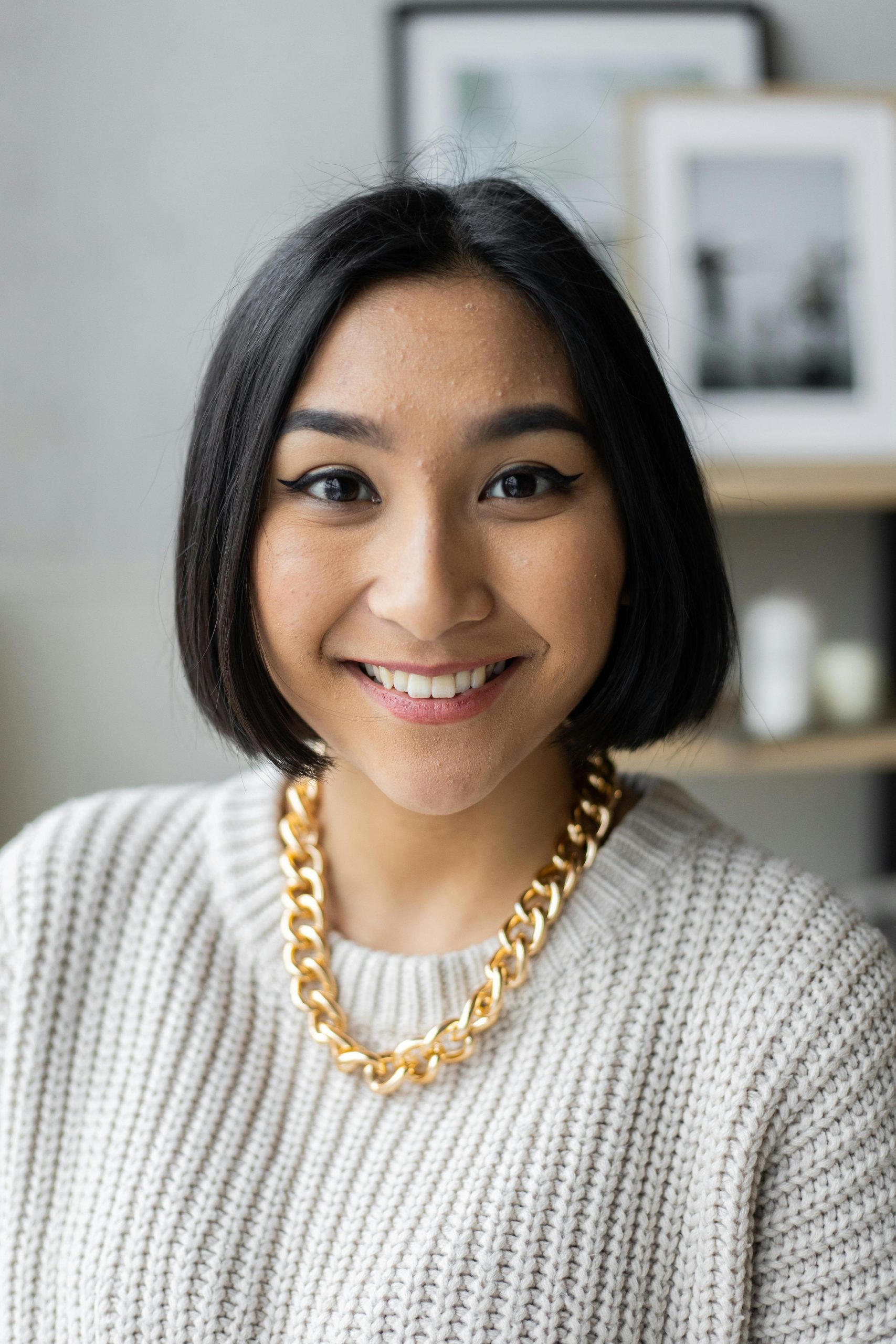 Positive young Asian female in knitted sweater with thick golden chain on neck against blurred background looking at camera